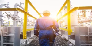Worker in blue overalls and a white hard hat walking through an industrial plant with extensive piping, representing tasks in oil and gas or chemical processing where non-sparking tools are essential for ATEX zone compliance.